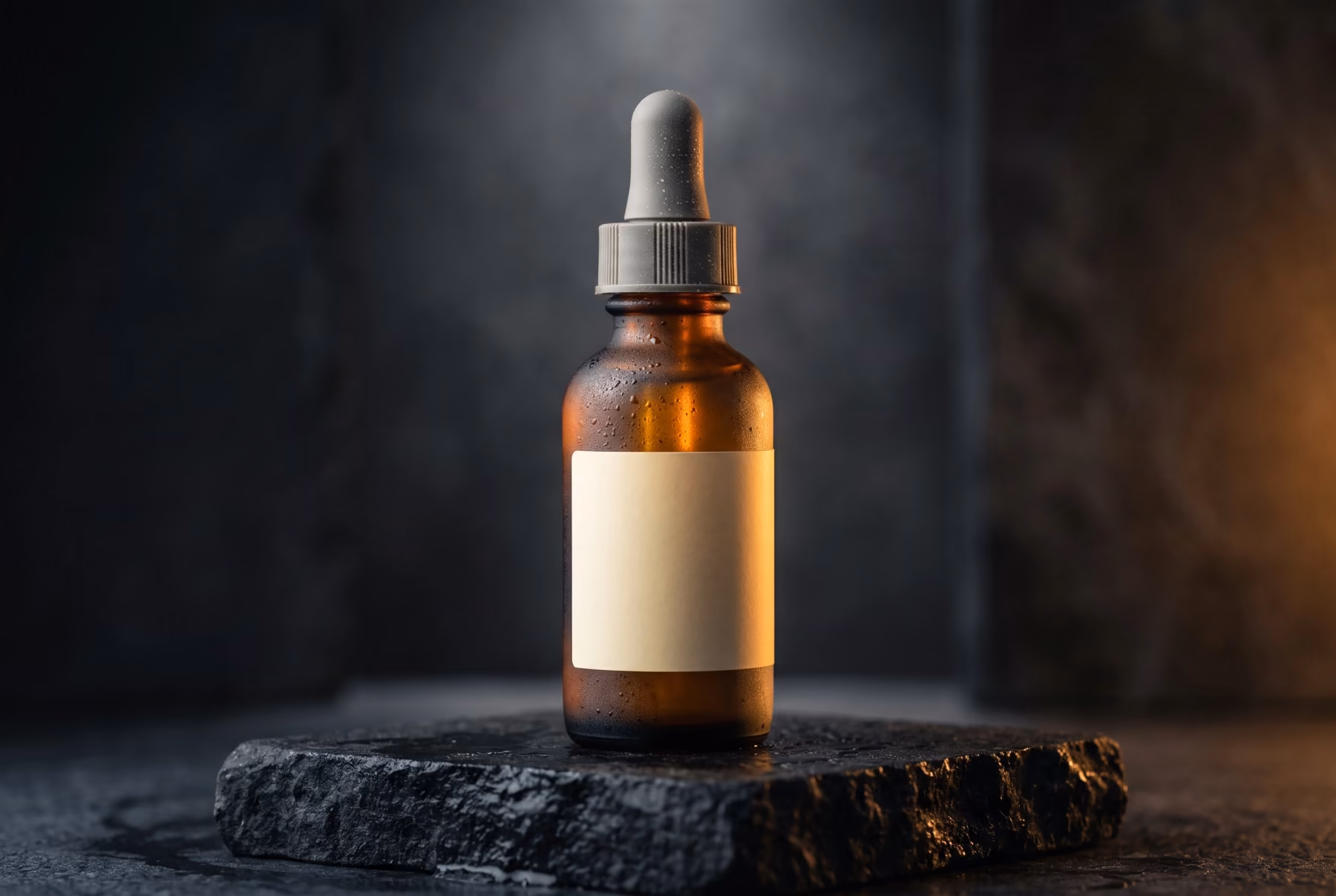A frosted amber glass serum bottle with a grey dropper cap standing on a dark stone plinth, backlit in a hazy studio.