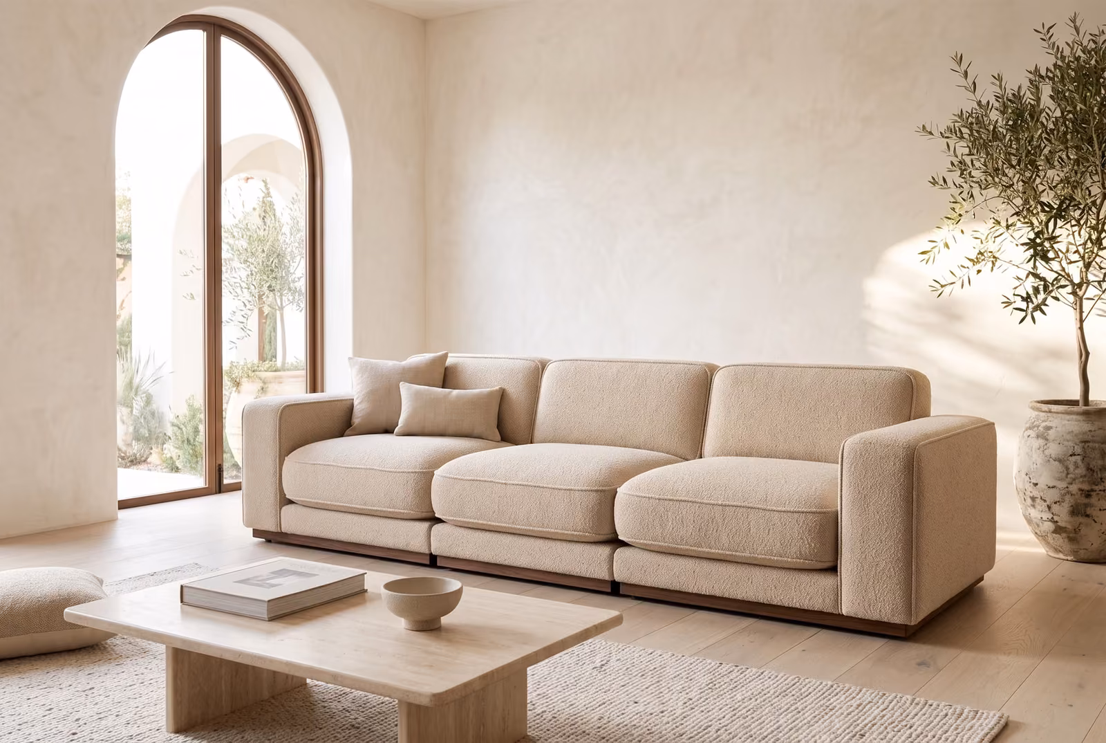 A low three-module sofa in oatmeal boucle on a walnut plinth, in a bright living room with an arched window, limewash walls, a travertine coffee table and an olive tree.