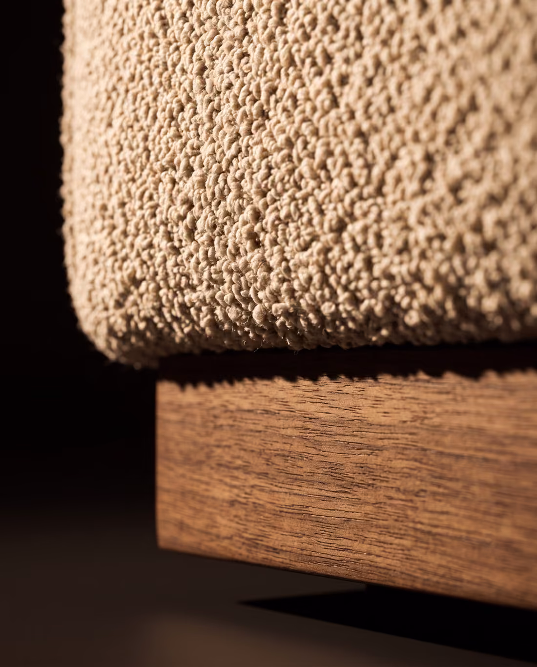 Close-up where the boucle upholstery overhangs the oiled walnut plinth, showing the wood's straight grain.