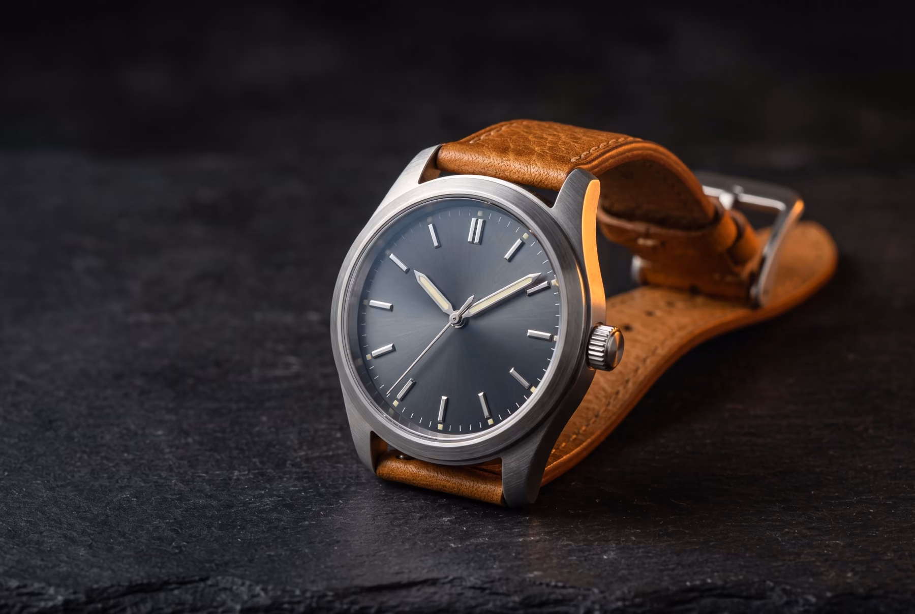 A field watch with a brushed steel case, slate dial and tan leather strap resting on dark stone under a warm rim light.