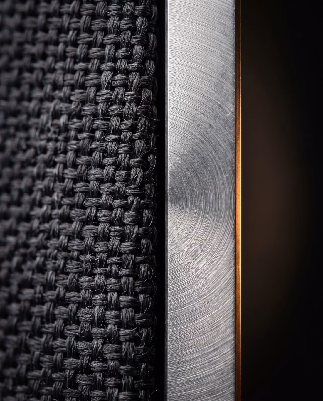 Extreme close-up of charcoal woven speaker fabric meeting a brushed aluminum seam.
