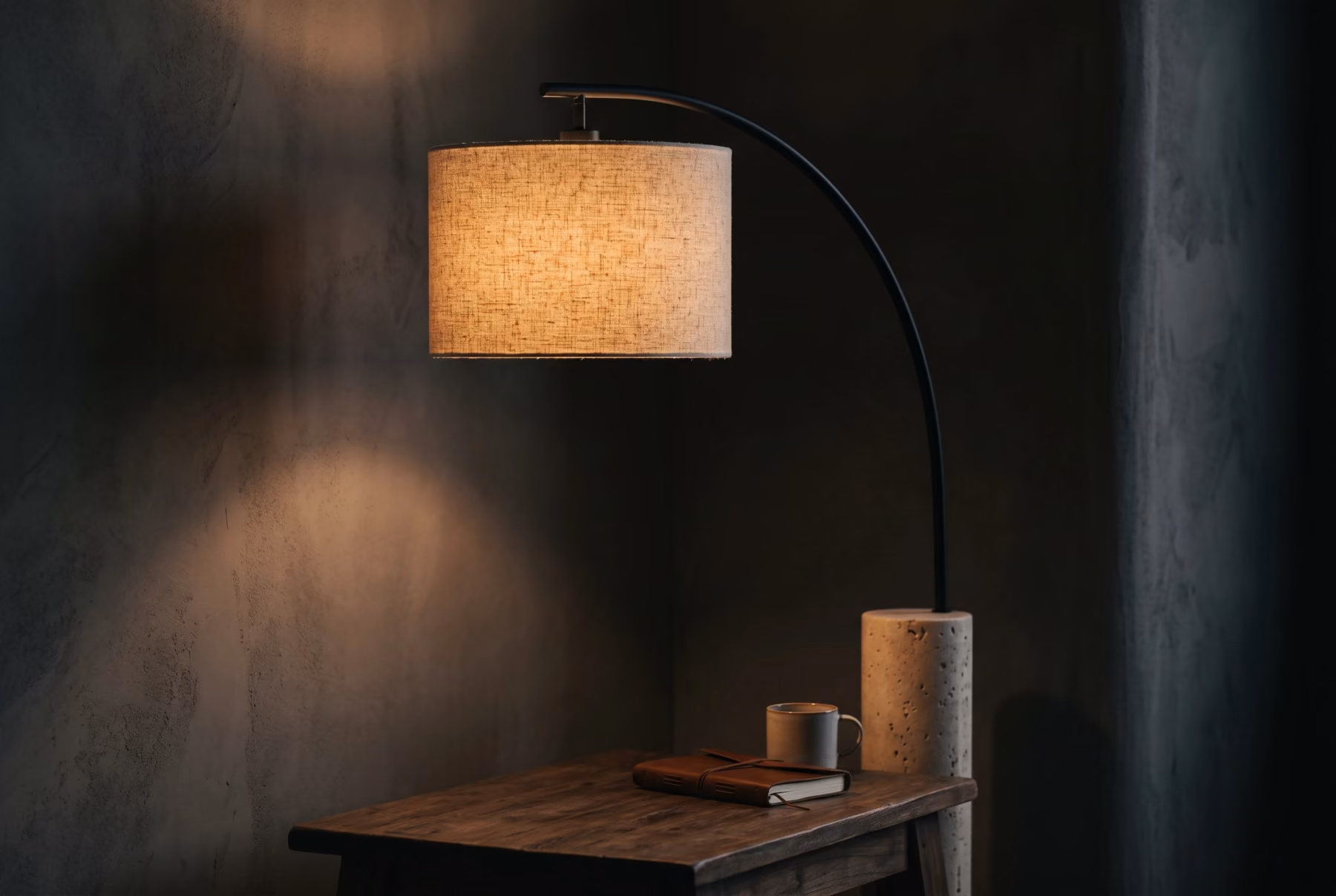 An arc table lamp with a blackened steel stem and warm linen shade glowing in a dark minimal interior corner.