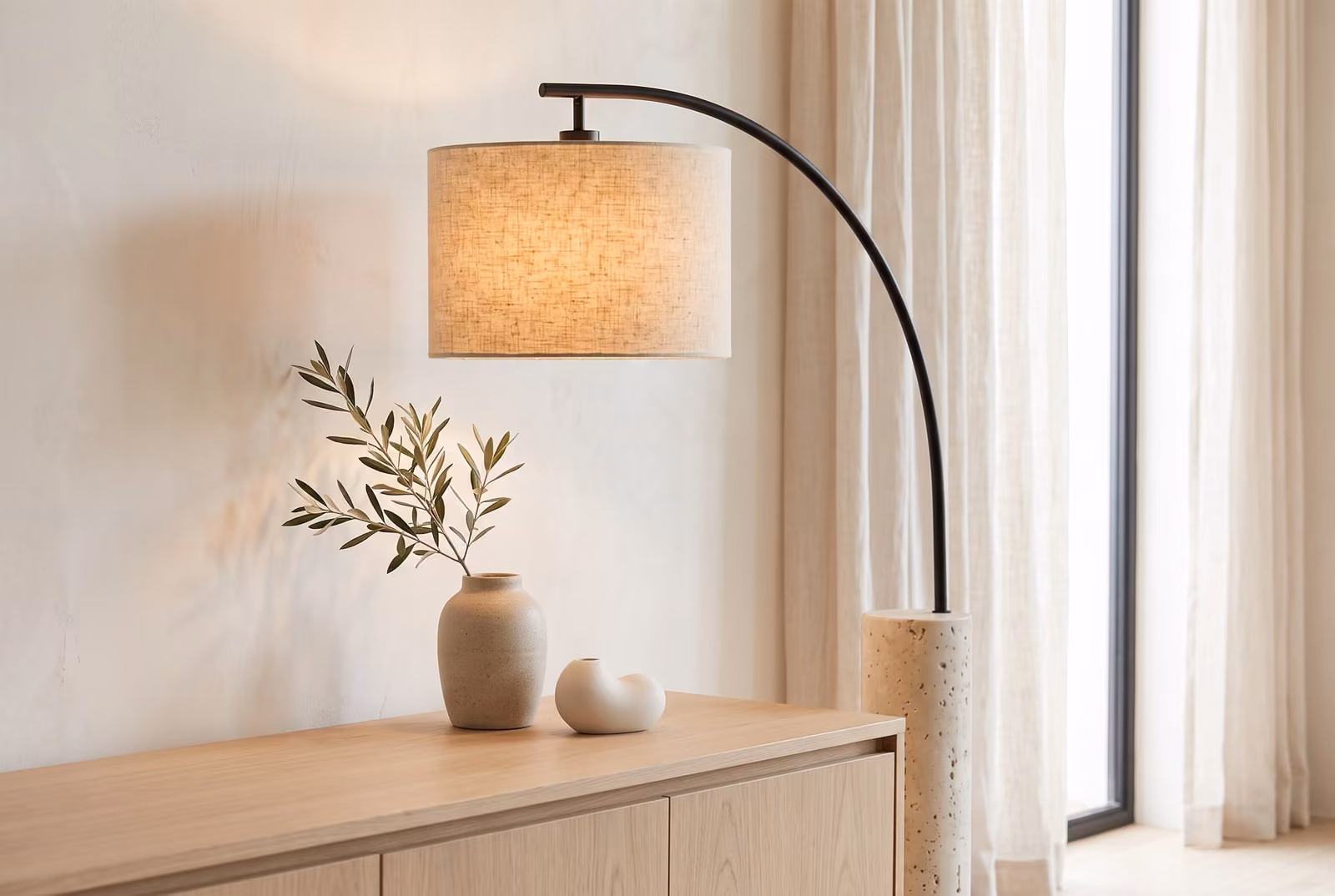 An arc floor lamp with a black steel arm, linen drum shade and travertine column base, beside an oak sideboard in a bright living room.