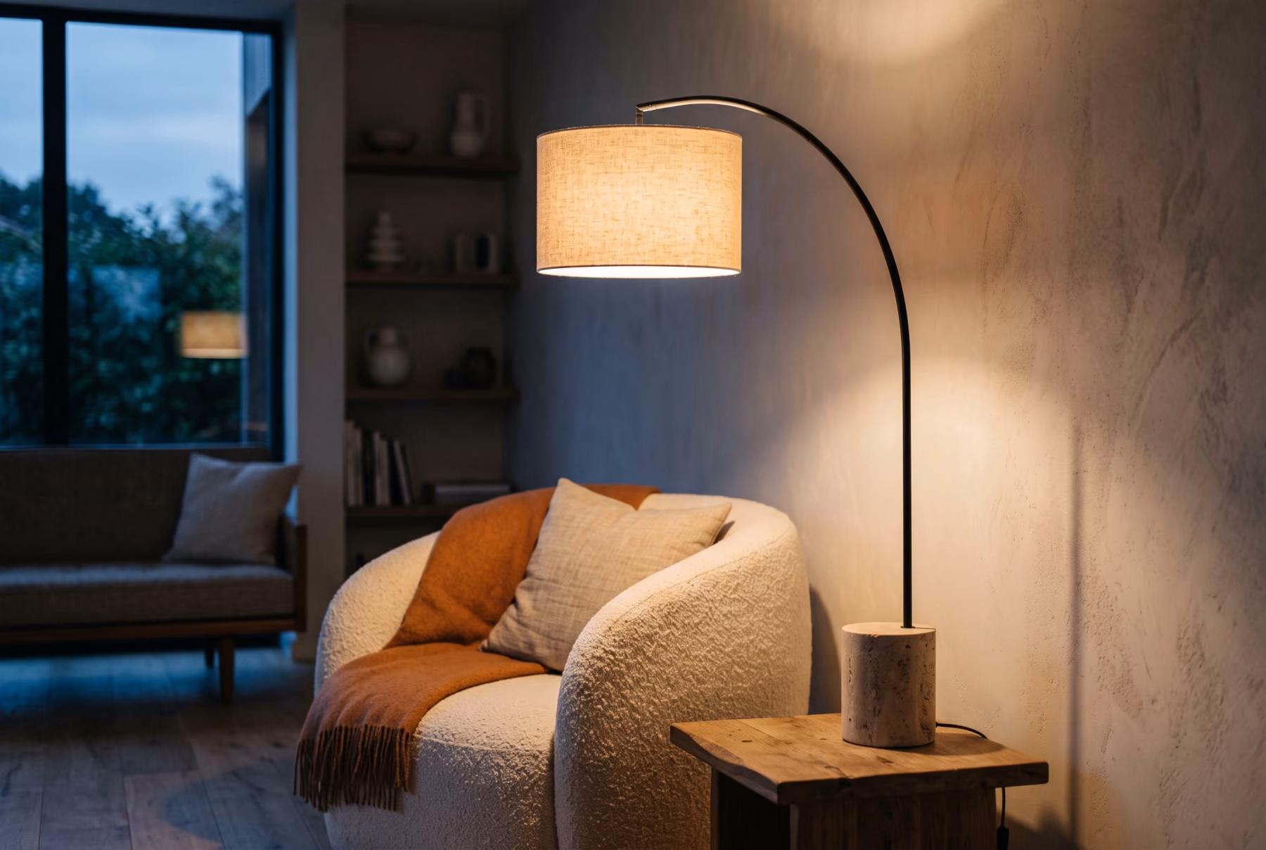 The lamp switched on beside a boucle armchair in a calm living room at dusk, casting a warm pool of light on the wall.