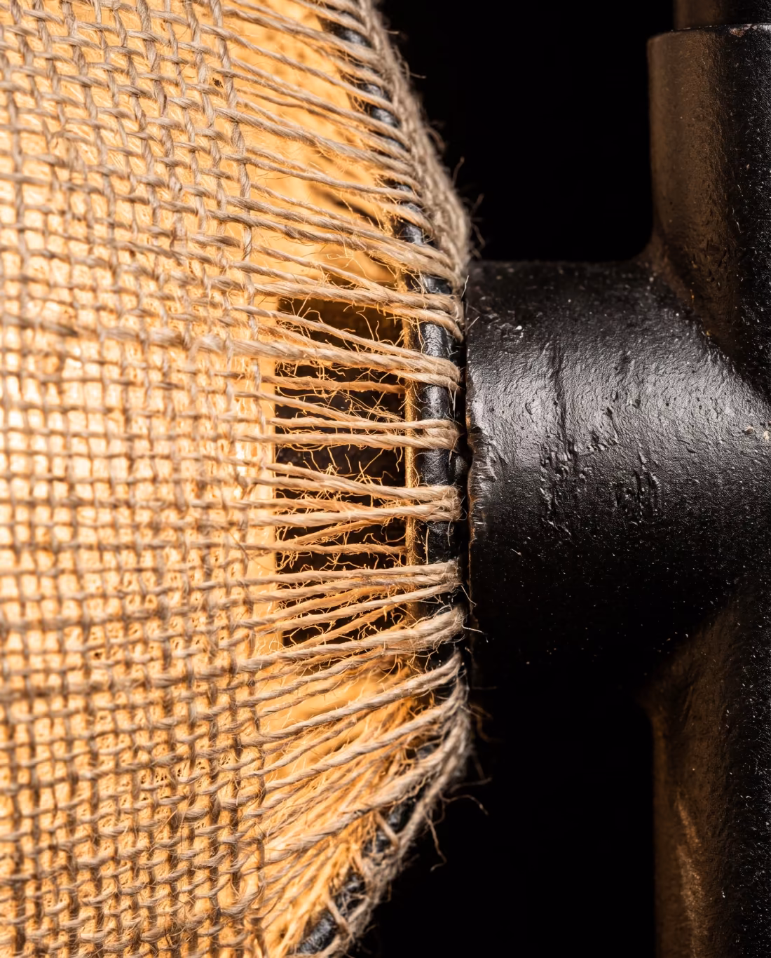 Extreme close-up of a warm linen lampshade weave lit from within, meeting a blackened steel fitting.