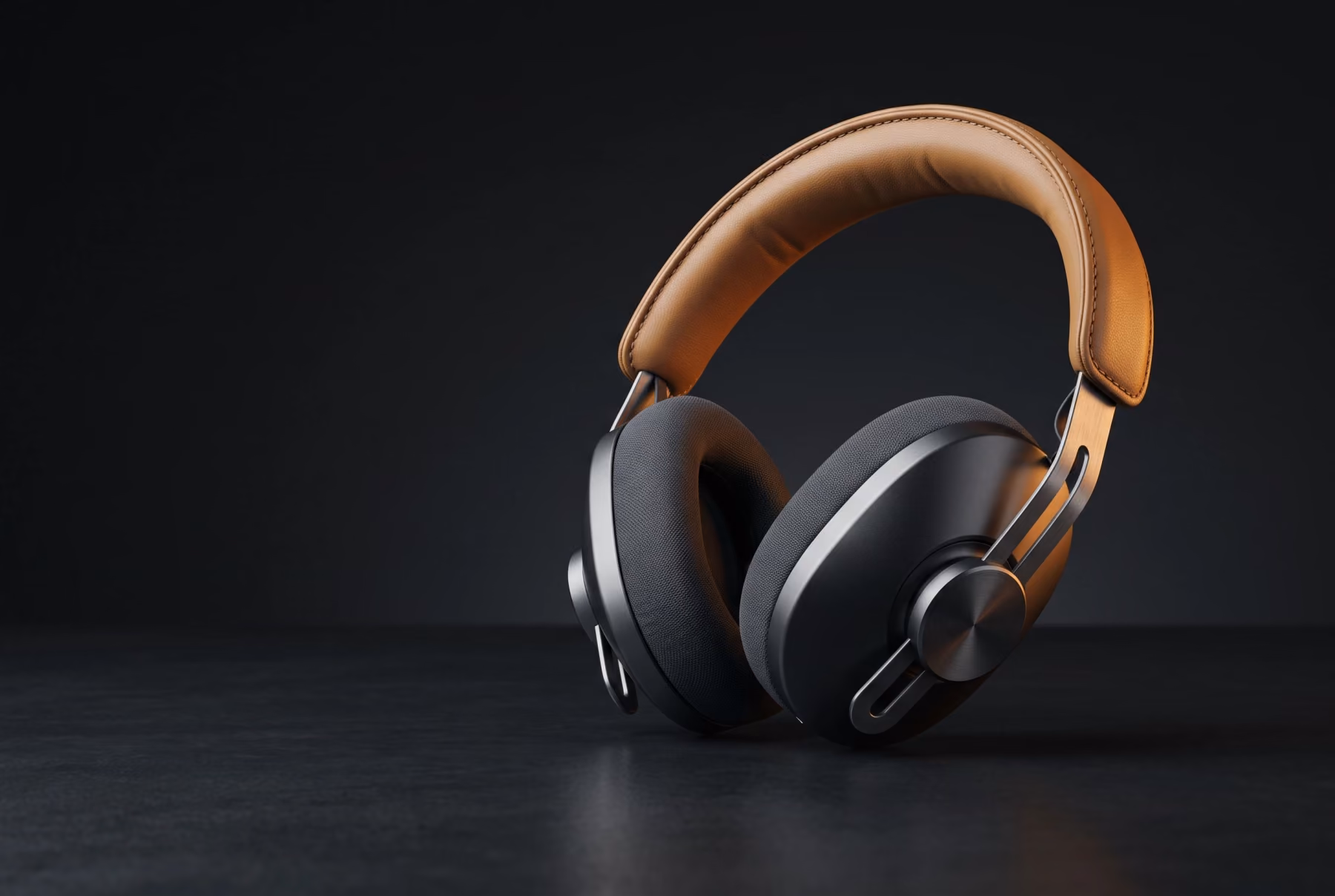 Matte charcoal over-ear headphones with a sand leather headband, resting at an angle in a dark studio under a warm amber rim light.